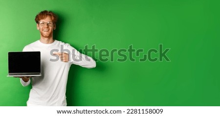 Happy redhead guy in glasses and white long-sleeve t-shirt, pointing finger at blank laptop screen and smiling, standing over green background.
