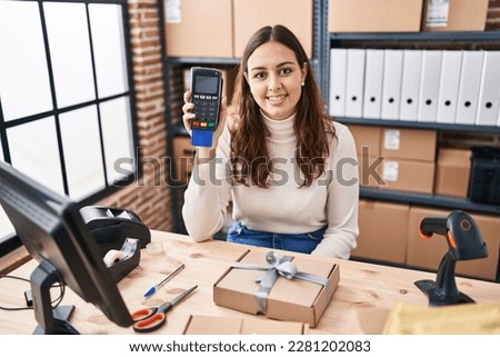 Young hispanic woman working at small business ecommerce holding dataphone looking positive and happy standing and smiling with a confident smile showing teeth 