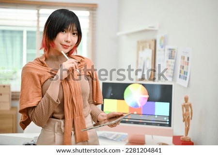 Portrait of a smiling and pretty young Asian female web graphic designer or interior designer stands in her studio with her digital tablet.