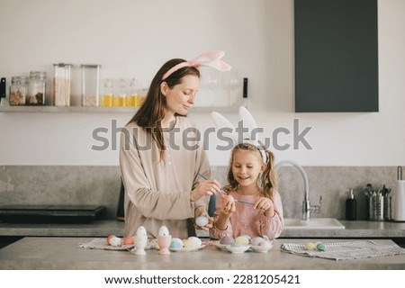 Happy mother and her little daugter decorating Easter eggs using brush and paint on the kitchen. Holiday preparations.