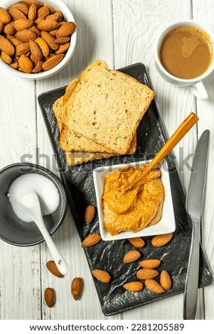 Raw Organic Almond Butter in a bowl on table with wooden spoon and almonds scattered on the table, toast bread and coffee. Top View