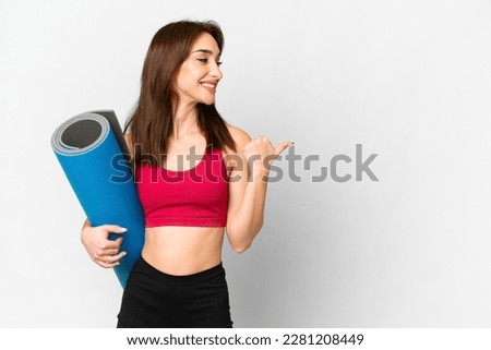 Young sport caucasian woman going to yoga classes while holding a mat over isolated white background pointing to the side to present a product