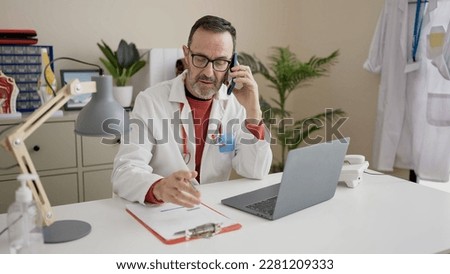 Middle age man doctor using laptop talking on smartphone at clinic