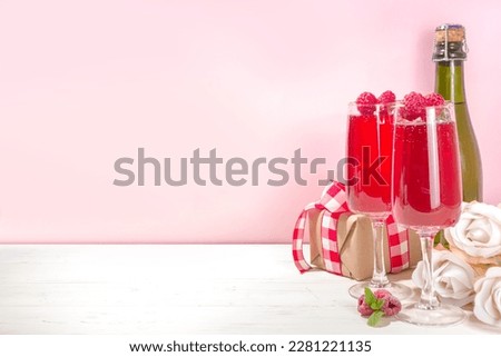 Red mimosas cocktail for Valentine's. Classic mimosa drink with raspberry juice, raspberry and mint decor. With gift box for Valentine day, rose flowers, champagne bottle