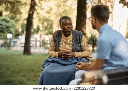 African American senior man in wheelchair and caregiver communicating while relaxing in the park of residential care home. Royalty-Free Stock Photo #2281228929