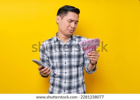 Portrait of crying young Asian man wearing plaid shirt holding mobile phone and cash money in rupiah banknotes isolated on yellow background. people lifestyle concept