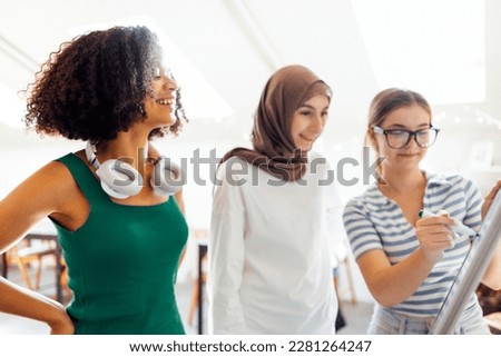 Brainstorming of three teenagers. Teens of different nationalities and religions solve problems together. White girl writes on blackboard with marker. Muslim and african american female are smiling.