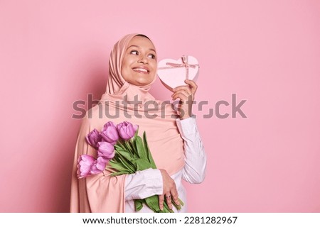 Happy Arab Muslim woman wearing pink hijab, holding a beautiful bouquet of purple tulips and a heart shaped gift box, smiling cutely looking aside, isolated over pink background with copy ad space