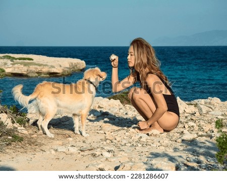 Model, of Asian appearance, sits on a cliff near the Mediterranean Sea. In a black one-piece swimsuit. She gives commands to the reddish dog Kokoni Royalty-Free Stock Photo #2281286607