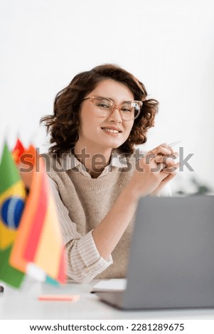 cheerful language teacher in glasses sitting next to laptop and flags on blurred foreground