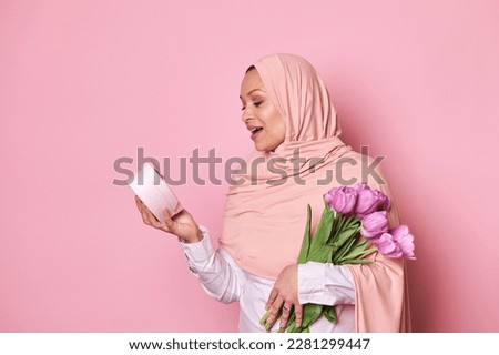 Surprised Muslim pretty woman in pink hijab, holding a bouquet of tulips, expressing amazement while getting a happy present in a pink gift box for festive occasion, isolated over pink background