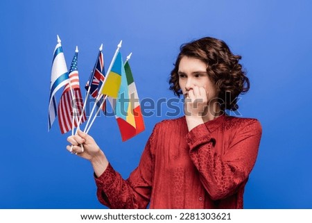thoughtful woman holding hand near face while looking at flags of different countries isolated on blue