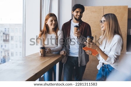 Group of young coworkers in casual clothes standing at table with cups of coffee and enjoying coffee break at window in daylight in modern office Royalty-Free Stock Photo #2281311583