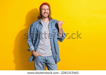 Photo of friendly guy with long hairdo dressed jeans shirt look directing empty space hand in pocket isolated on yellow color background