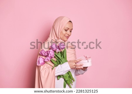 Middle-aged smiling positive Muslim woman wearing a pink hijab, holding a bouquet of tulips and cute gift box for birthday, Women's or Mother's Day, isolated on pink background. Copy space for ads