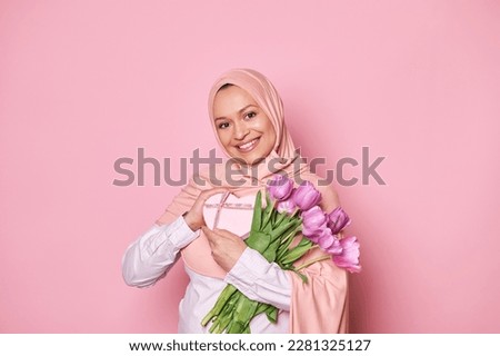 Positive Middle-Eastern Muslim woman in pink hijab with Mother's Day, birthday present or for any festive occasion and bouquet of purple tulips, smiling looking at camera, on isolated pink background