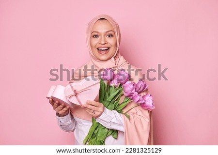Cheerful Arab lady in pink hijab, holding a heart shaped gift box and tulips, smiling to camera. Positive amazed Muslim woman with Mother's Day or birthday present posing over pink studio background
