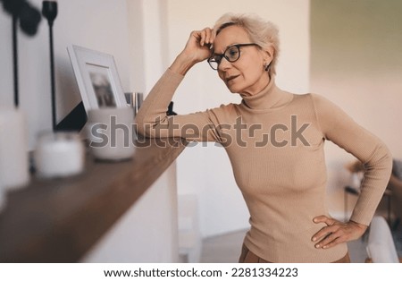 Exalted slim aged lady wearing casual clothes leaning on shelf at home while reminiscing past years and looking at old photo in frame