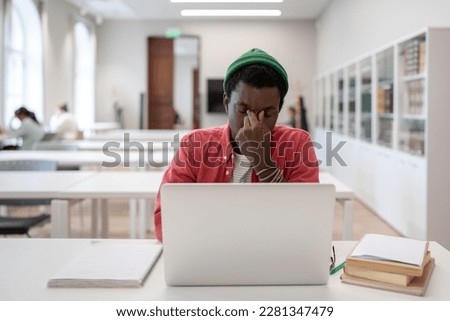 Academic stress. Tired frustrated black guy sitting in front of laptop in university library, suffering from digital eye strain caused by online education. Computer vision syndrome in students Royalty-Free Stock Photo #2281347479