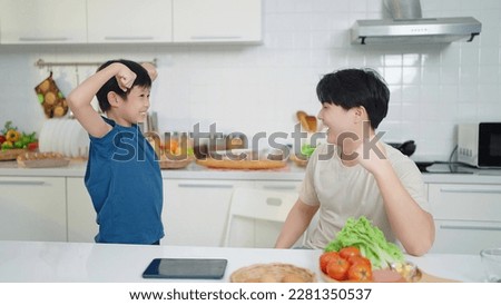Asian little cute boy in blue shirt demonstrating his muscles or biceps to father after eating food at kitchen room. Boy power concept , Happy family , Single dad Royalty-Free Stock Photo #2281350537
