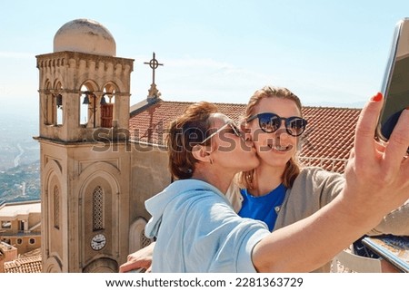 Young traveler girlfriends have fun and enjoy sunny day in mountain town using smartphone to take selfie with antique cityscape in background while hugging and kissing. Travel concept.