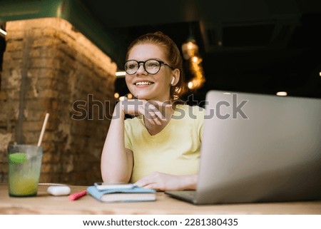 Cheerful adult female freelancer in casual clothes and eyeglasses browsing laptop while sitting at table with glass of lemonade and listening to song
