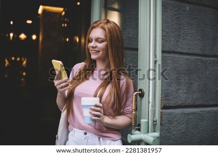 Positive young female standing with cup of hot beverage at cafe entrance door and smiling while looking at screen of mobile phone and passing time in daylight on street