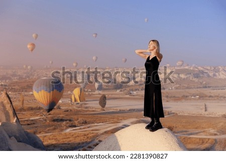 Traveler woman in black dress background landscape Cappadocia with hot air balloons sun light. Concept trip Turkey travel.