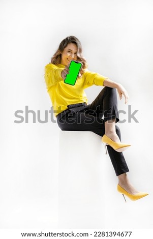 A young woman is sitting on a bedside table and shows a smartphone with a green screen.