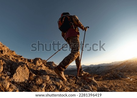 Hiker with backpack walk uphill in mountains. Active tourist silhouette goes with hiking poles at sunset