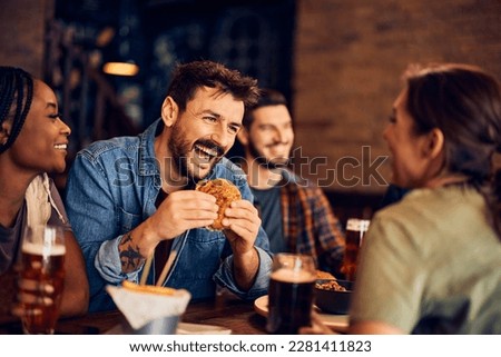 Cheerful man eating burger and having fun while gathering with friends in a bar.  Royalty-Free Stock Photo #2281411823