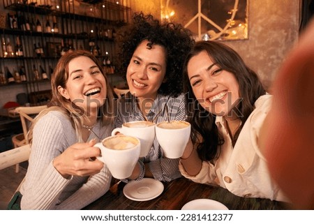 Group of cheerful female friends take a selfie toasting cups of cafe in coffee shop. Smiling and happy young girls looking at camera and having fun together. Women active on social networks.