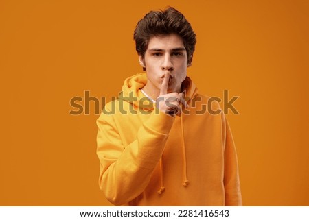 Young man hushing at camera and telling to be quiet on yellow background