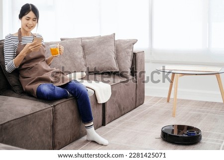 Black robotic vacuum cleaner cleaning the floor while woman sitting on sofa and relax.Young woman using automatic vacuum cleaner to clean the floor, controlling smart machine housework robot.