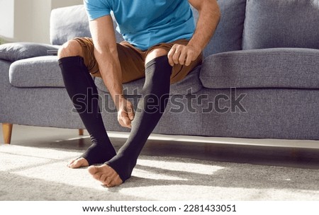 Cropped photo of a man putting on black medical compression stockings on his legs for the prevention of varicose veins and for venouse therapy sitting on the couch at home. Healthcare concept. Royalty-Free Stock Photo #2281433051