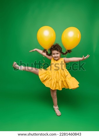 Funny little girl on a green background. A girl is having fun with yellow balloons. balloons lift up the pigtails of the child.