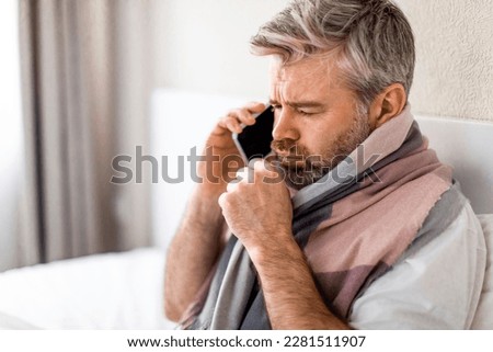 Sad sick middle aged grey-haired man with warm scarf around his neck sitting in bed at home, talking the phone to his employer while asking about the sick leave, calling doctor, coughing, copy space Royalty-Free Stock Photo #2281511907