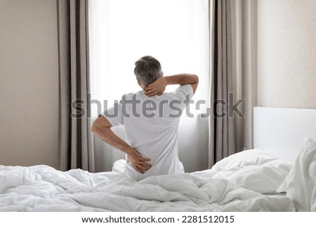 Unrecognizable mature grey-haired man wearing white pajamas sittting on bed at home in the morning, touching lower back and neck, suffering from muscle strain after waking up, copy space Royalty-Free Stock Photo #2281512015