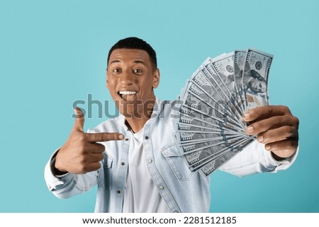 Glad millennial black male in casual shows finger to many dollars, rejoices in victory online, recommends savings isolated on blue studio background. Win and finance, profit money Royalty-Free Stock Photo #2281512185