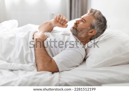 Chronic cough, reasons and treatment. Unhealthy sick grey-haired bearded middle aged man wearing pajamas lying in bed and coughing, home interior, side view, copy space Royalty-Free Stock Photo #2281512199