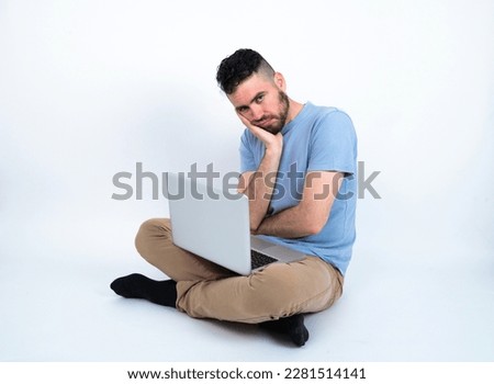 Very bored Young caucasian man with laptop sitting over white studio holding hand on cheek while support it with another crossed hand, looking tired and sick,