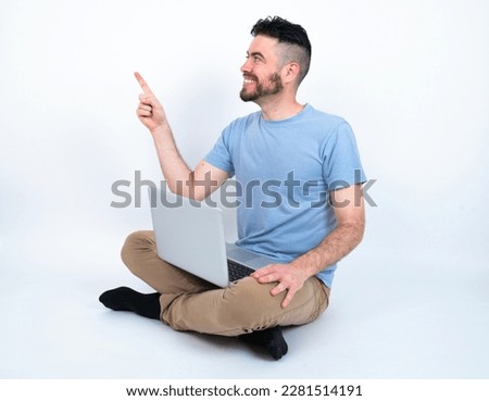 Smiling Young caucasian man with laptop sitting over white studio indicating finger empty space showing best low prices