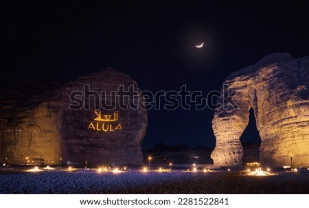 Jabal AlFil - Elephant Rock in evening, landscape illuminated, text Alula and Arabic translation projected to stone wall seats for people set up on the ground, crescent moon - composite image - above Royalty-Free Stock Photo #2281522841