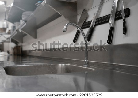 Metal shiny chrome kitchen counter with sink and faucet under knives on wall in modern restaurant