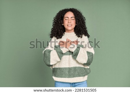 Young calm happy latin woman holding hands on chest isolated on green background. Smiling pleased grateful female model expressing gratitude, mental balance, feeling love and warmth concept.