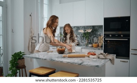 Cute family cooking at kitchen together. Young woman and child girl preparing dough on table. Mother daughter sifting flour in modern interior house. Adorable kid helping parent at light white home