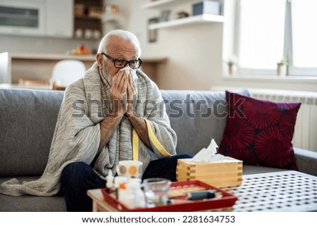 Senior man suffering from flu while sitting wrapped in a blanket on the sofa at home. Elderly Man blowing his nose while lying sick in bed at home. Man with a cold lying in sofa holding tissues Royalty-Free Stock Photo #2281634753