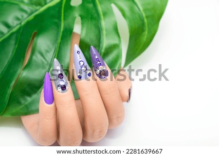 Female hand with vacation stiletto nail design. Glitter purple and blue nail polish manicure with rhinestones and glitter nail art. Female model hand hold tropic leaf on white background. Copy space Royalty-Free Stock Photo #2281639667