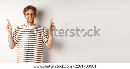 Funny young redhead guy in nerdy glasses pointing fingers sideways, showing two promo offers, standing happy over white background.