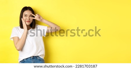 Lovely asian girl in white t-shirt posing with hand on cheek, showing peace sign on eye, standing over yellow background.